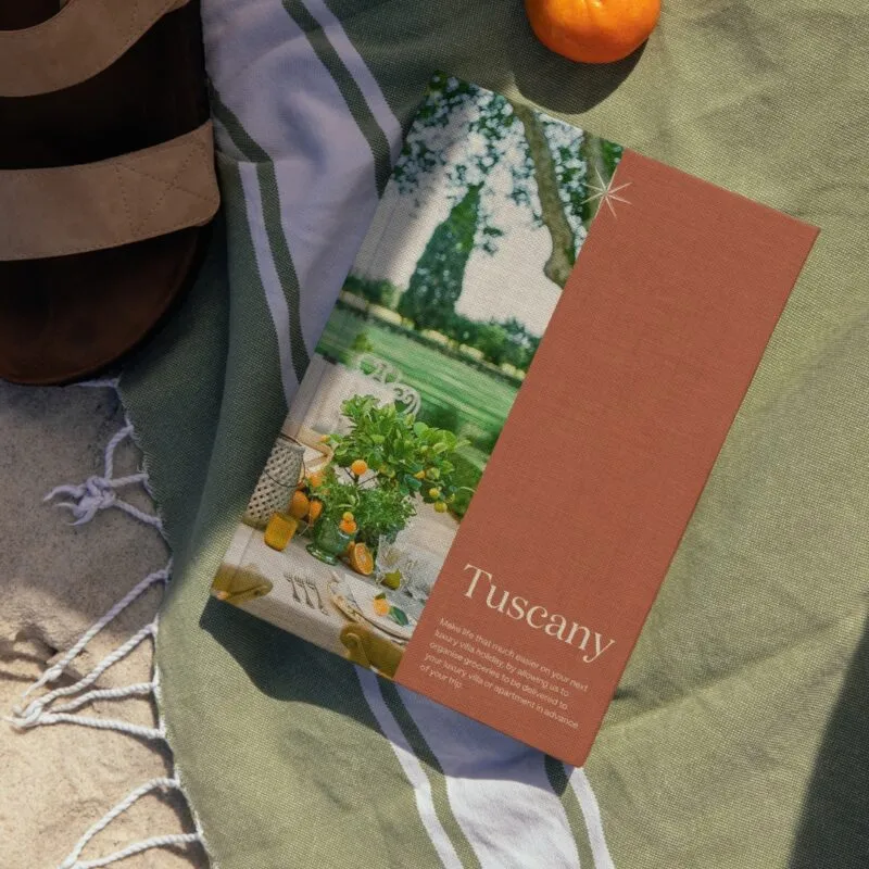 In relaxing, sandy scenery, a book rests casually on a soft pistachio-coloured blanket with white stripes, next to a pair of stylish sandals and a tangerine. The book's cover is visible – divided vertically in the middle, its left side is occupied by a beautiful photograph of a Tuscan countryside villa, with greenery, trees and outdoor furniture visible; the right side of the book cover is rusty orange. The bottom of that side is occupied by the title of the book and a short description, both in white. The title, in large headline text, says 'Tuscany', and underneath it, in smaller copy text, the description reads 'Make life that much easier on your next luxury villa holiday, by allowing us to organise groceries to be delivered to your luxury villa or apartment in advance of your trip.'. The elegant white compass marque of The Luxury Travel Book decorates the book in top centre, it's North and South indicators aligning perfectly with the photo/colour divide on the book cover, which is beautifully textured, almost as if it was made of canvas.