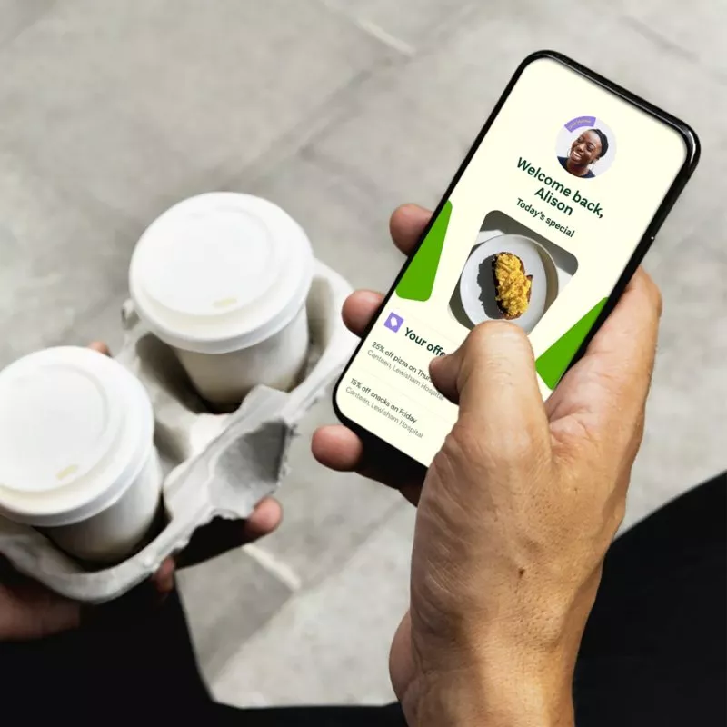 Image of mans hand holding phone with app screen , scrolling food options with personal profile top of screen. Holding coffee in the other hand
